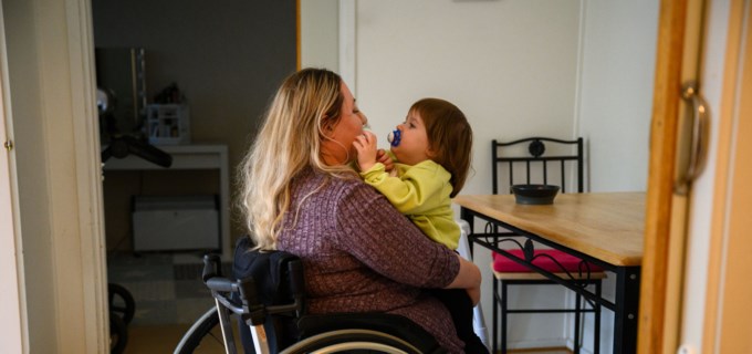 En mamma som sitter i rullstol och har ett barn i knäet. Barnet och mamman kramas och tittar på varandra. 