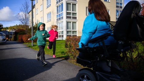 En person i blå jacka och med långt brunt hår kör en elektrisk rullstol på en asfaltsväg. Två glada barn kommer springande mot personen i rullstolen 