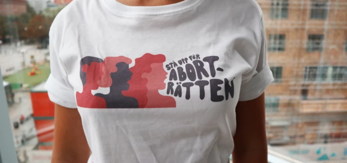 En person har på sig en vit t-shirt med en illustration av tre personer och texten "Stå upp för aborträtten". 