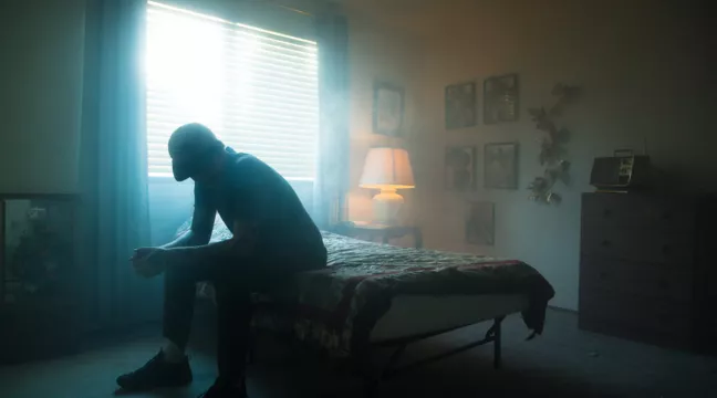 En person sitter på sängkanten i ett dunkelt rum 