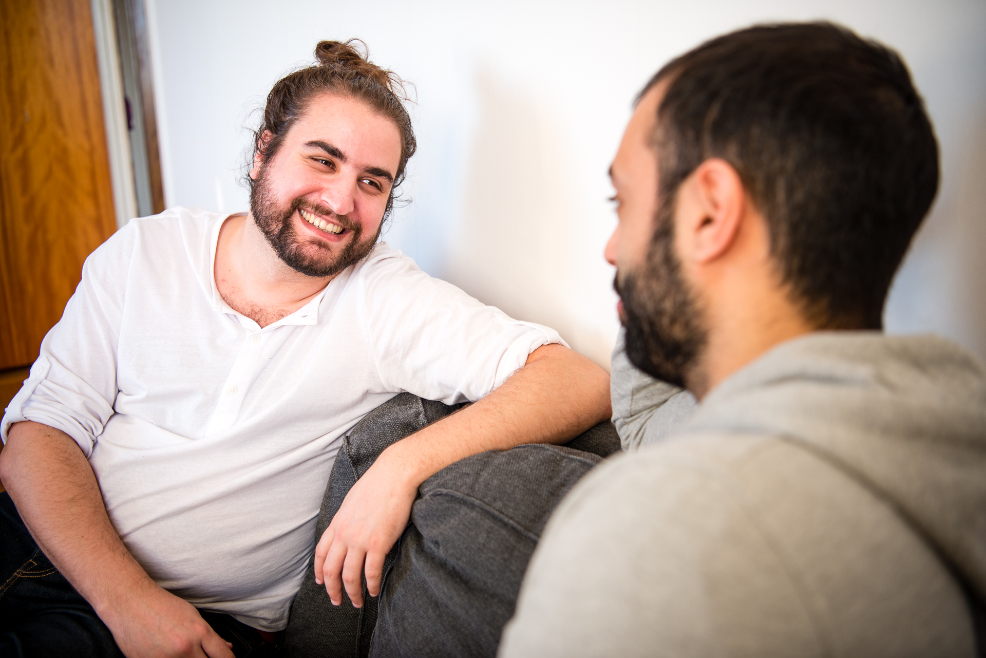 Två killar sitter i en soffa, håller varandra i handen, tittar varandra i ögonen och ler. 