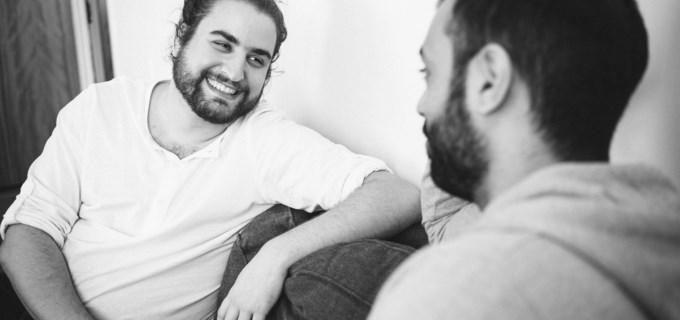 Två killar sitter i en soffa, håller varandra i handen, tittar varandra i ögonen och ler. 