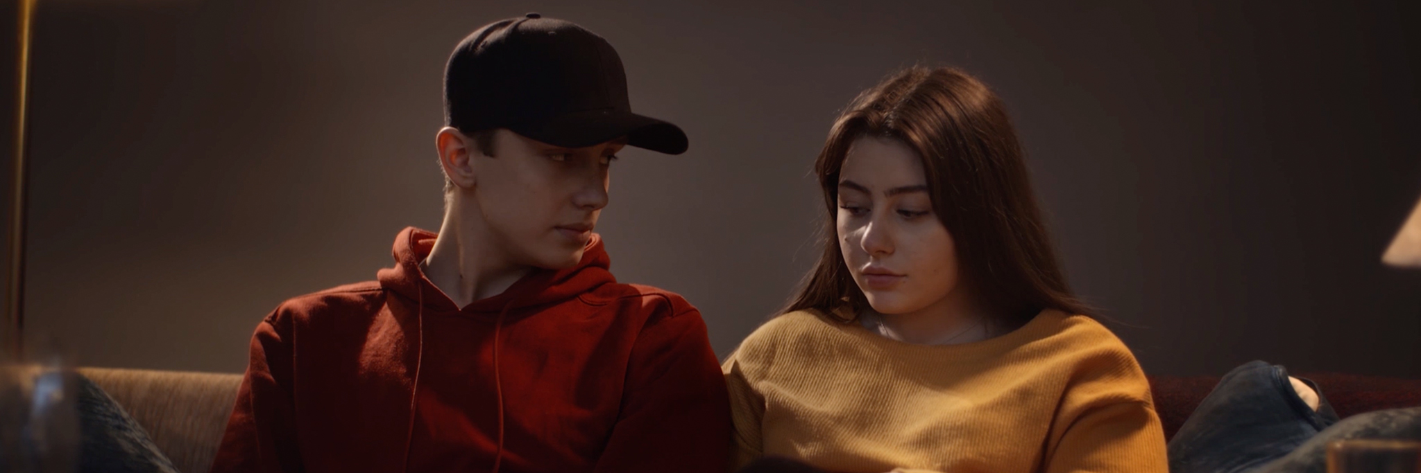 Två personer sitter i en soffa i dämpad belysning. Den ena tittar på den andra, som själv tittar ner. 