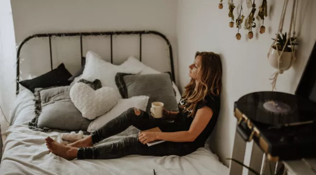 En kvinna sitter på sin säng med en kopp te i handen. Hon ser lite trött ut. 