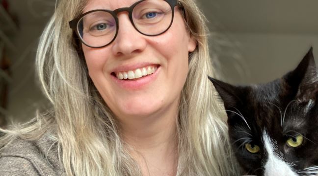 Blond person med glasögon ler och gosar med en katt. 