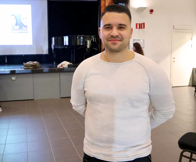 Person i vit tröja med händerna på ryggen tittar in i kameran.  