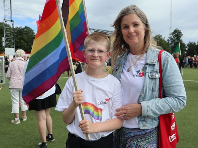 Ett barn och en vuxen med regnbågsflaggor tittar in i kameran och ser bestämda ut. 