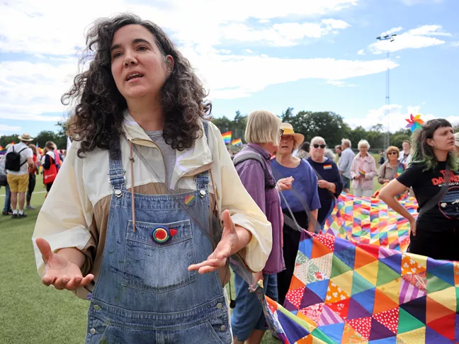 En person med snickarbyxor och lockigt hår gestikulerar. Bakom henne ett långt lapptäcke. 