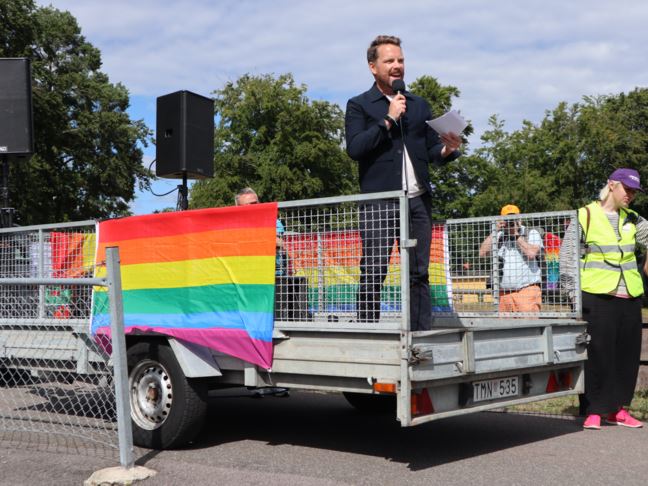 En person står och håller tal på en liten lastbil med regnbågsflagga på sidan. 