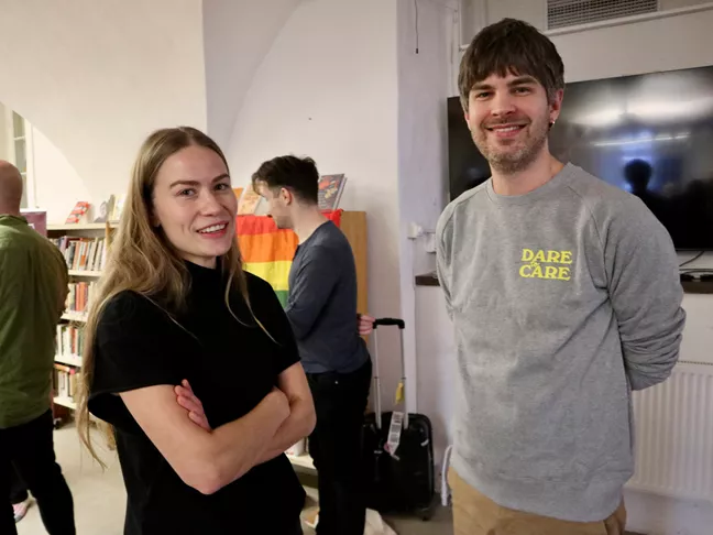 Två personer står bredvid varandra och tittar in i kameran. Den ena har långt hår och står med armarna i kors. Den andra har en tröja som det står Dare to Care på. 