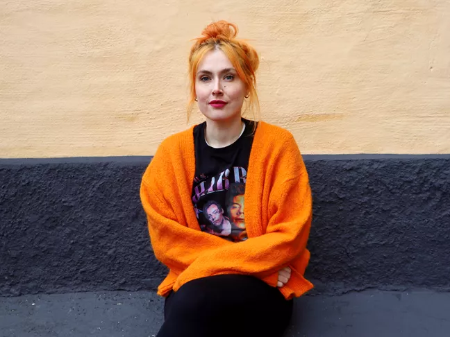 Person i rött hår, orange kofta och en t-shirt med Harry Styles sitter på en bänk. 