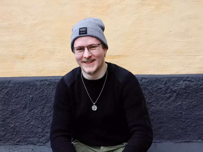 Person i svart tröja och grå mössa ler och ser lite busig ut. 