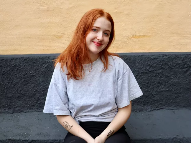 Person i grå t-shirt och med rött, långt hår ler och tittar in i kameran. 