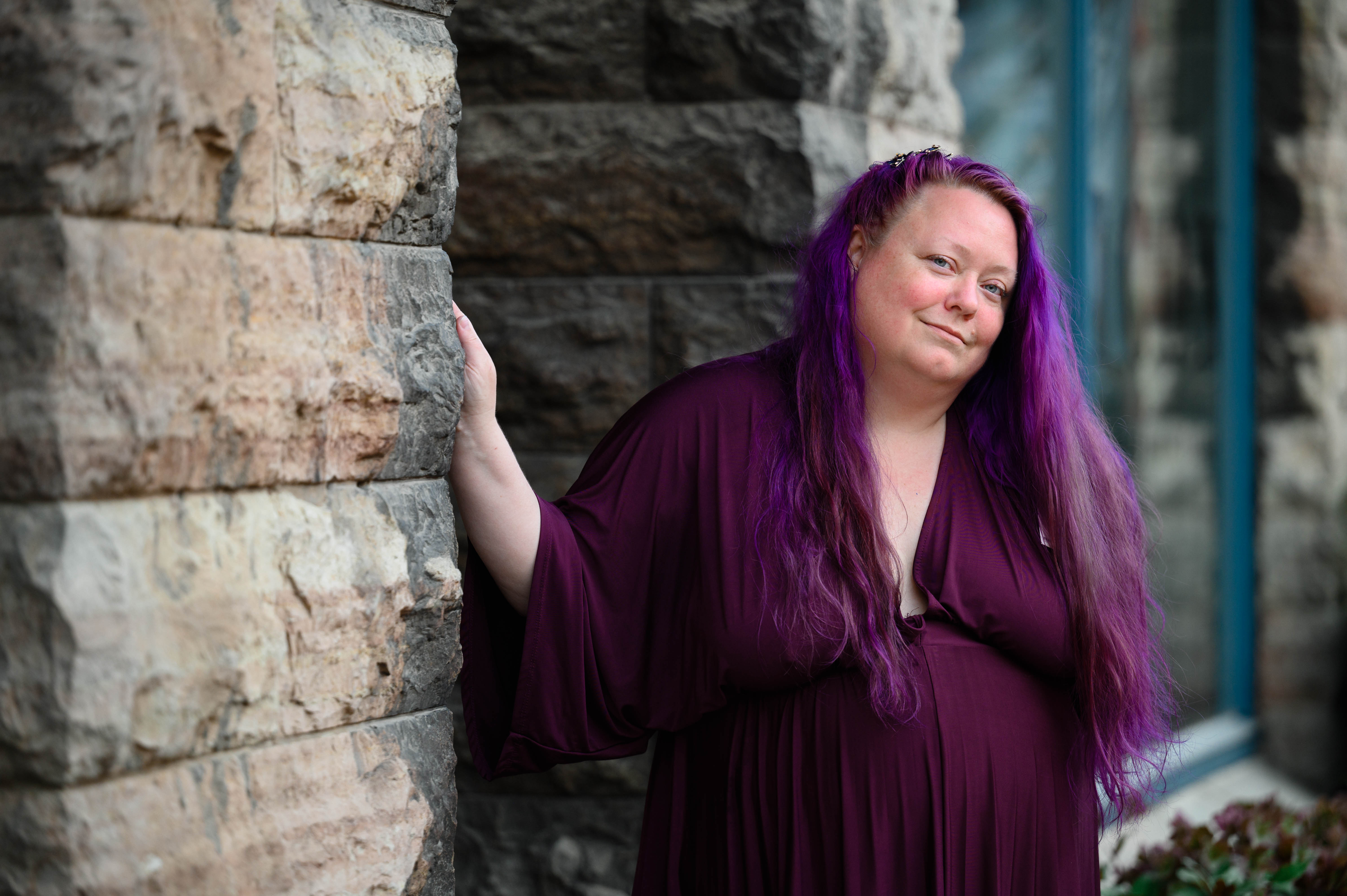 Lila står lutad mot en stenvägg. Hon har lila kläder och lila hår.  