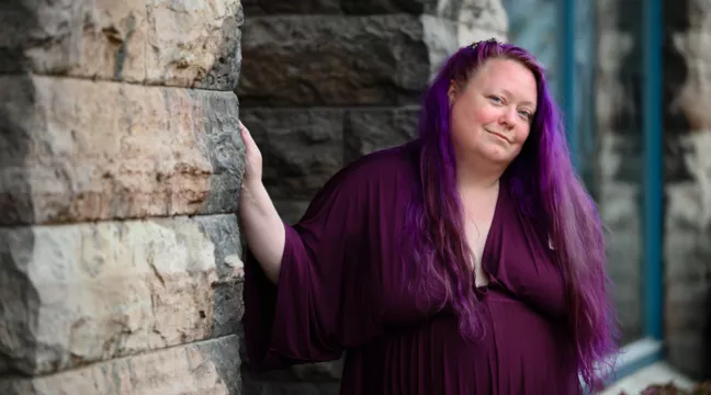 Lila står lutad mot en stenvägg. Hon har lila kläder och lila hår.  
