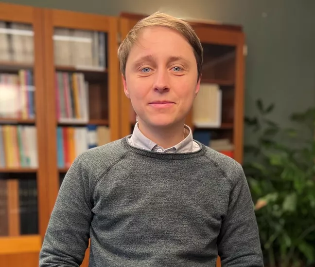 Jacko står i grå tröja och tittar in i kameran. I bakgrundens syns en bokhylla. 