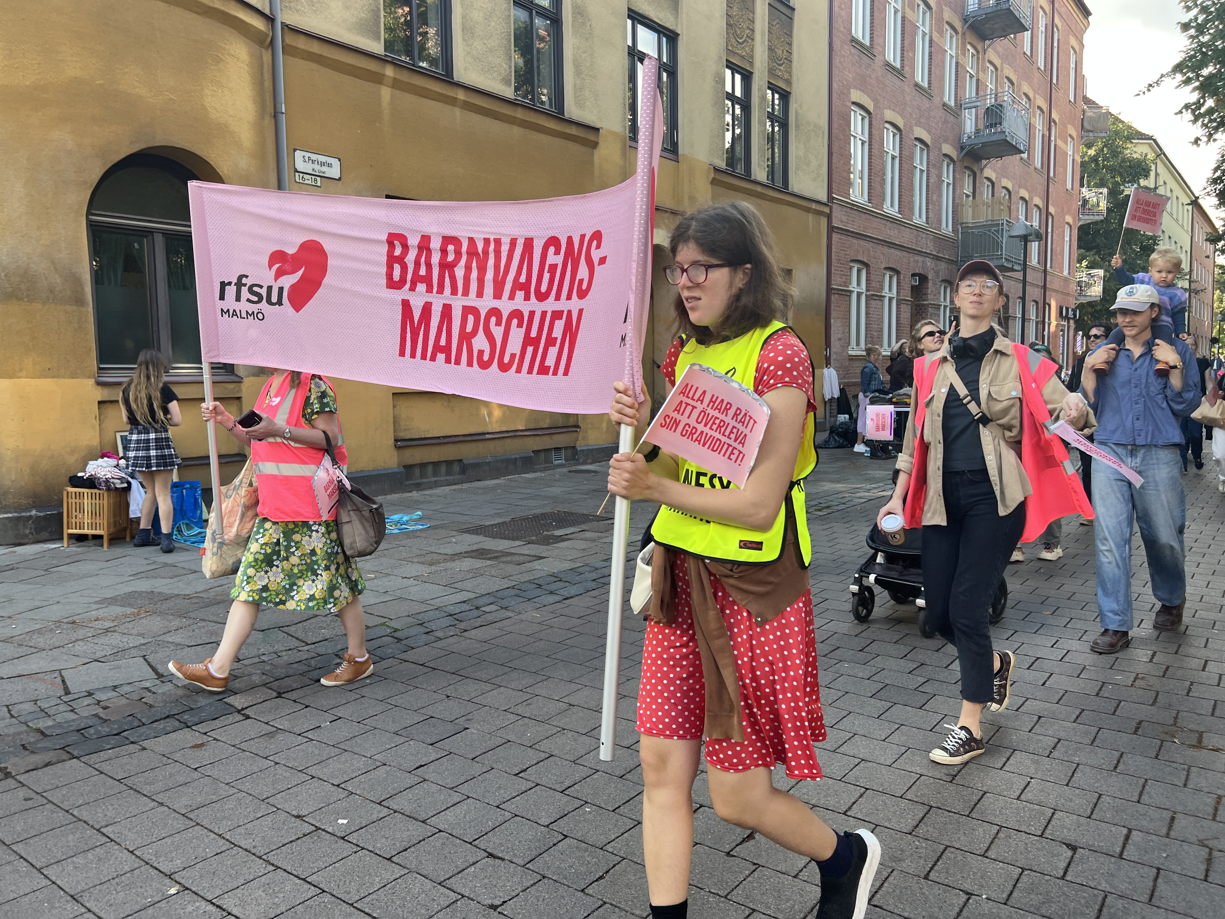 Två personer håller en stor banderoll som det står Barnvagnsmarschen på. De går på en gata med flera personer efter sig. 