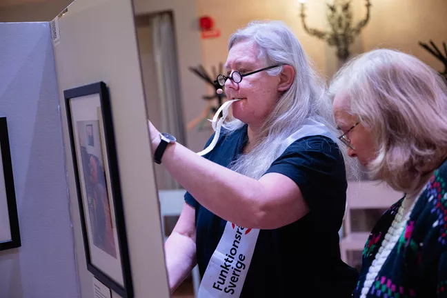 En person med långt grått hår har tejpbitar i munnen. Hon ser ut att klistra fast något på en pappskärm. 