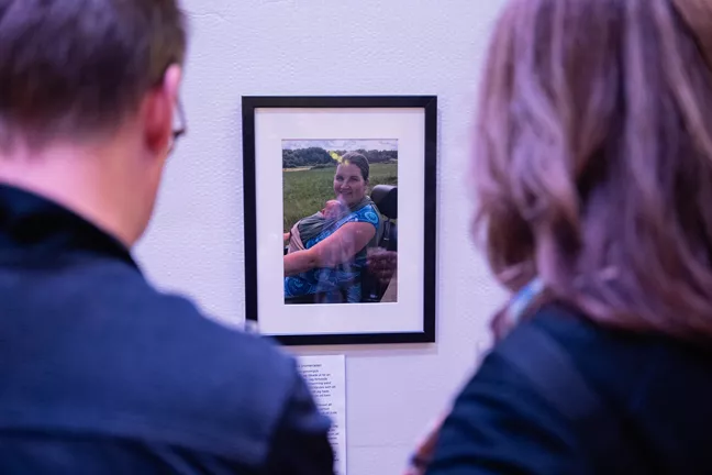 Två personer i förgrunden tittar på ett foto av en kvinna som sitter i rullstol med en bebis i bärsele. 