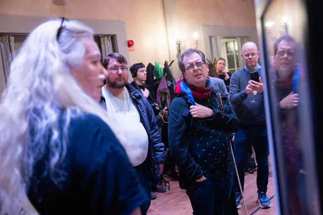 En person med glasögon tittar mot ett foto på väggen. Han har en käpp under ena armen. En kvinna i långt grått hår ser ut att berätta något för honom. 