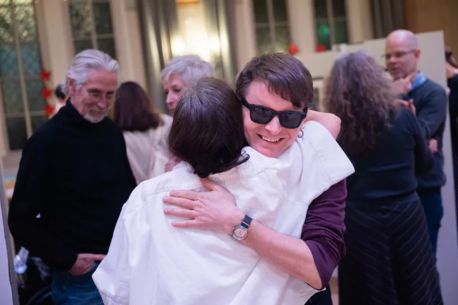 Två personer kramas. Den ena har mörka solglasögon. Den andra står med ryggen mot kameran. 