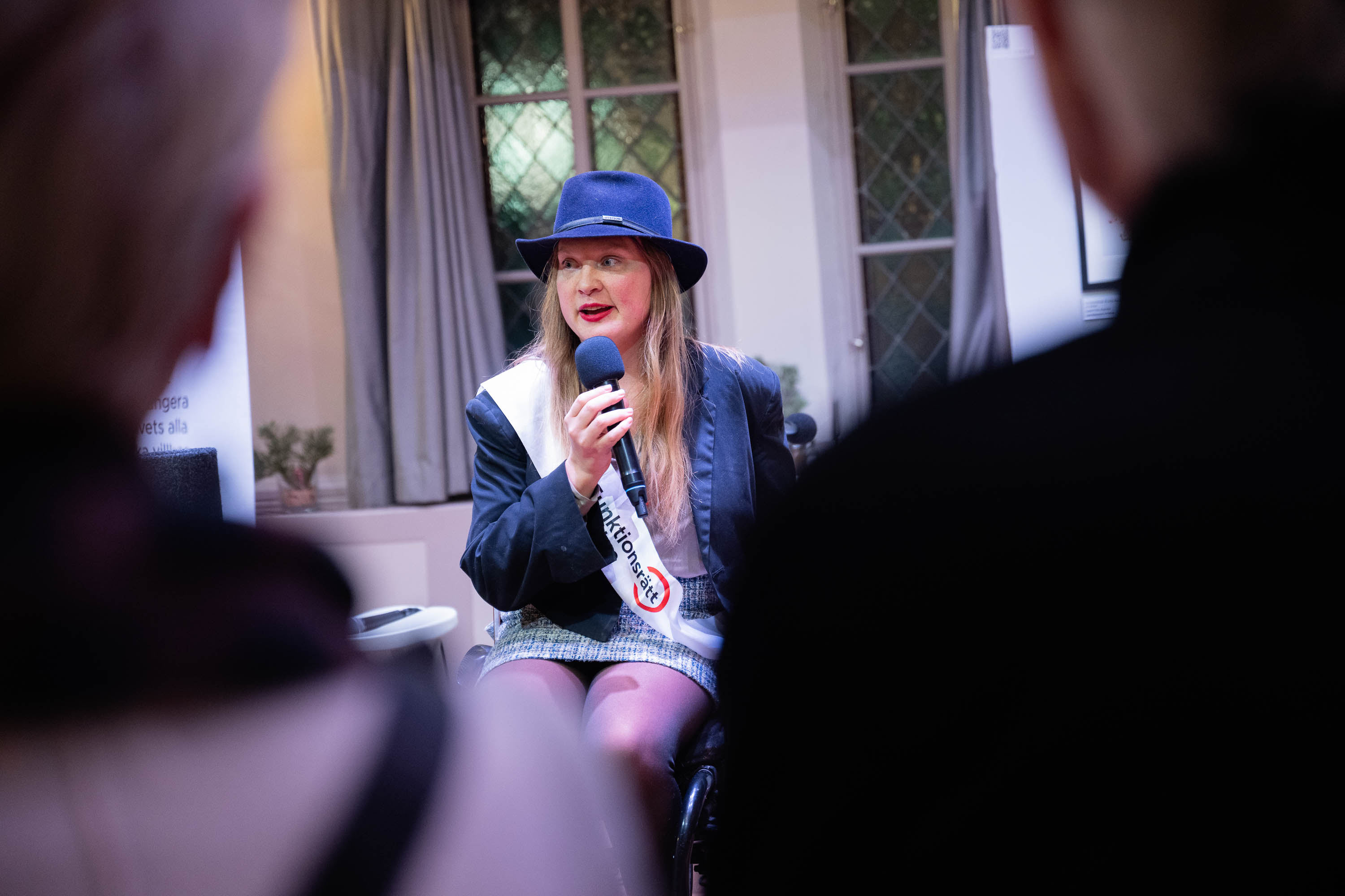 En person i hatt sittandes i rullstol pratar i mikrofon från en scen. 