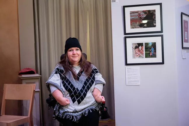 En person i svart mössa och långt hår sitter i en rullstol. Hon tittar in i kameran och ler. 