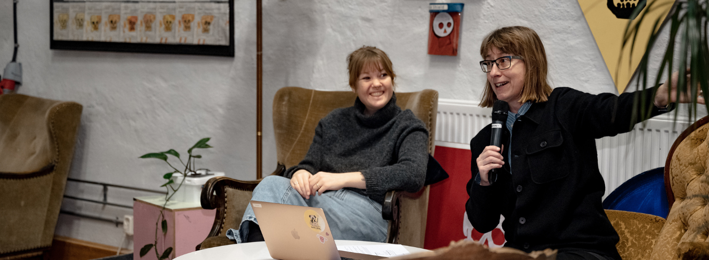 Två personer pratar inför publik. En ler åt den andre som pratar i mikrofon. 