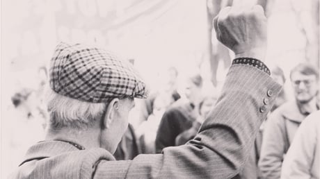Ett svartvitt fotografi taget bakifrån av en person i gubbkeps som höjer en knuten näve i luften framför en folksamling.
