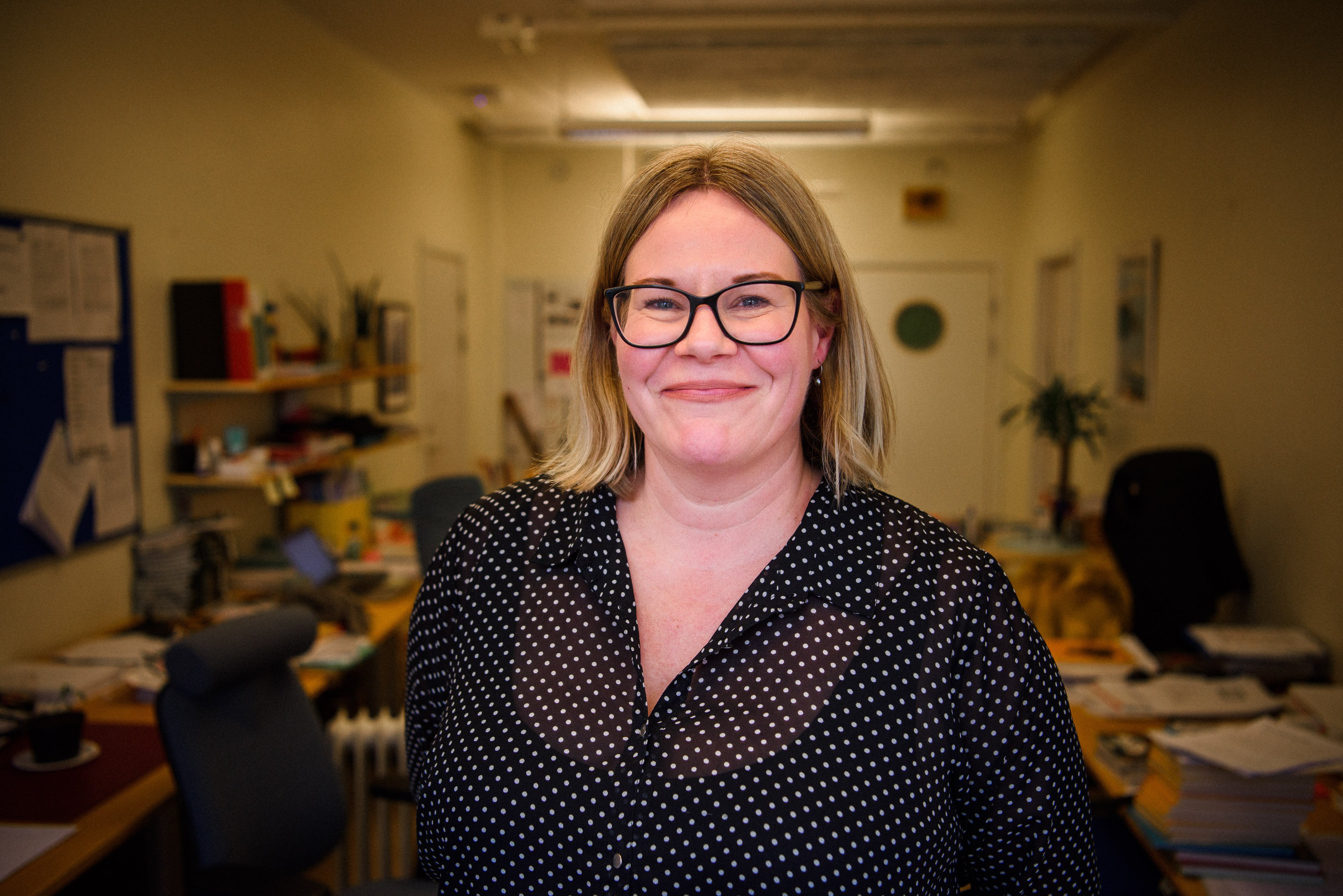 En person med glasögon och prickig blus ser in i kameran och ler. 