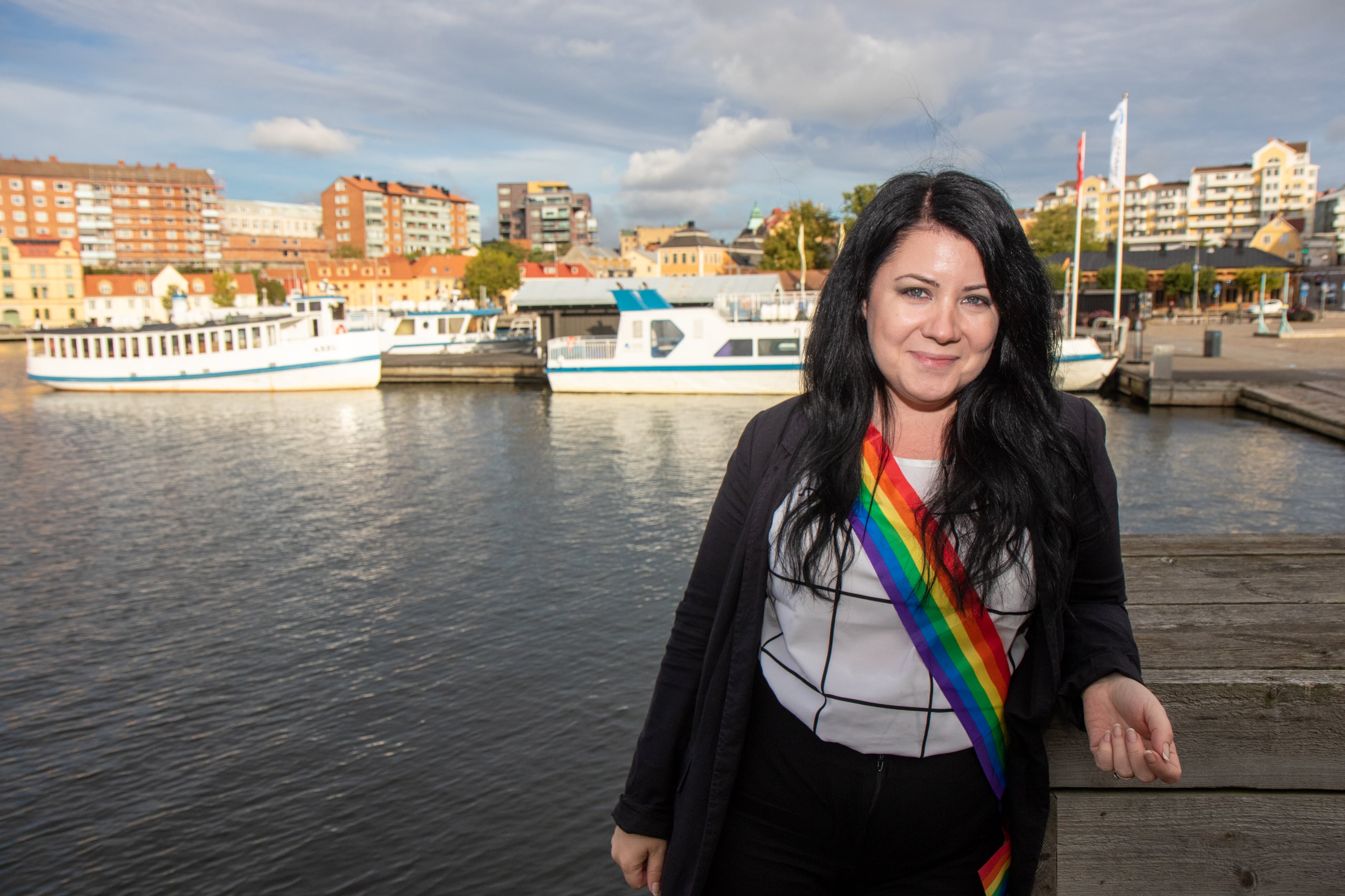 Sophia Ahlin står vid ett hamninlopp med båtar i bakgrunden och bär ett regnbågsfärgat ordensband över axeln.