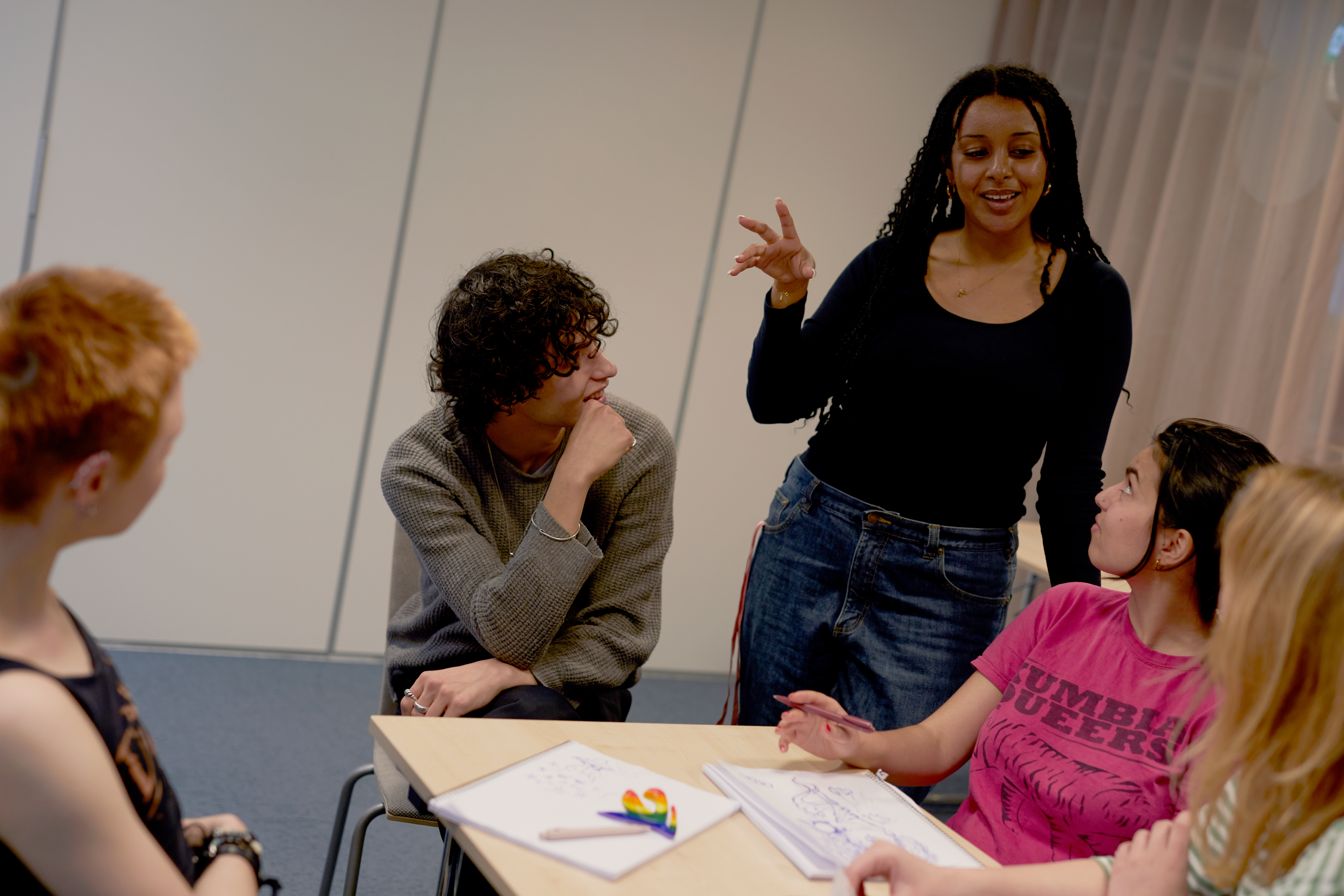 Woman is standing gesturing with her hands. A group of youths are sitting at a desk looking at her.