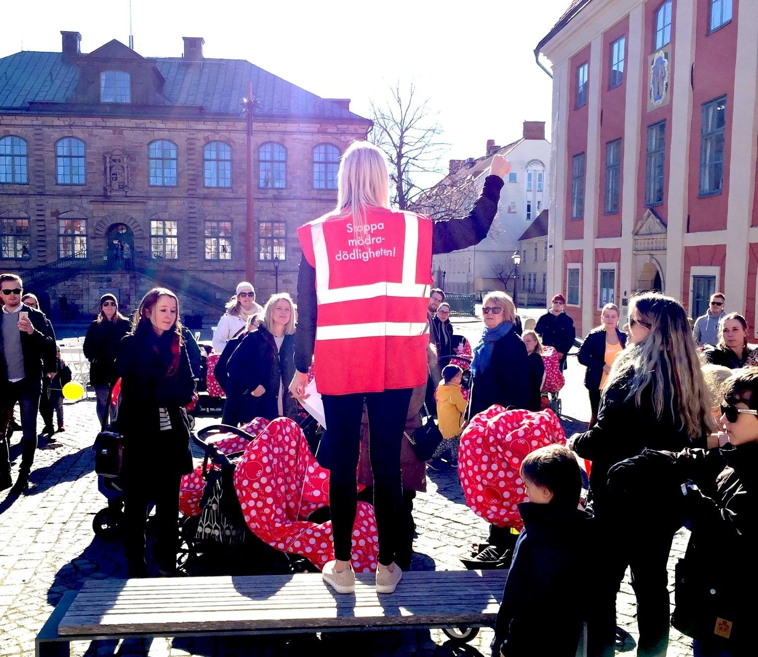 En person med röd väst står på en platå med handen upp i en knytnäve och pratar mot människor som står runt omkring med barnvagnar. På västen står det Stoppa mödradödligheten.