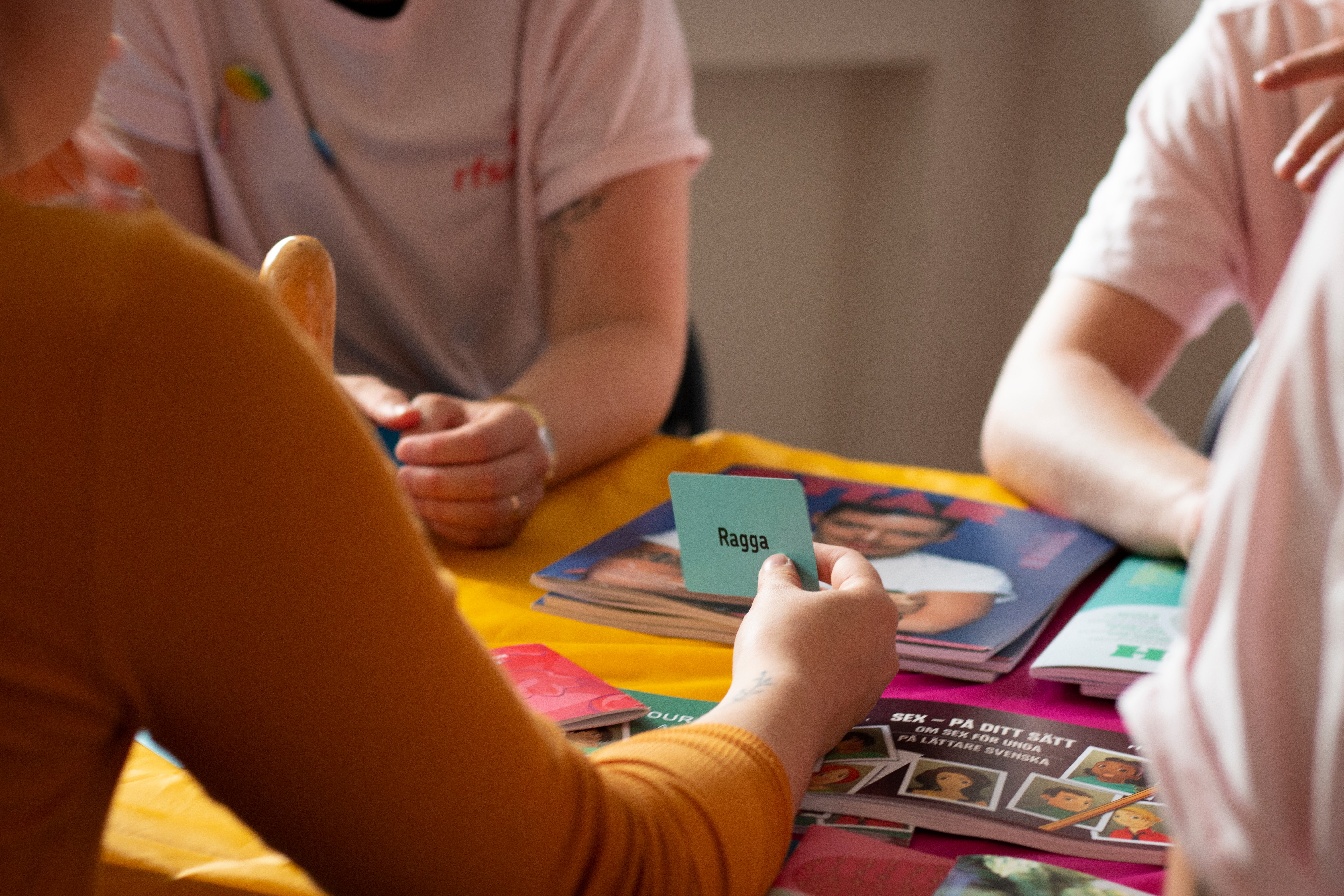 Fyra personer sitter vid ett bord och spelar ett kortspel. Endast händerna och överkropparna syns i bild.