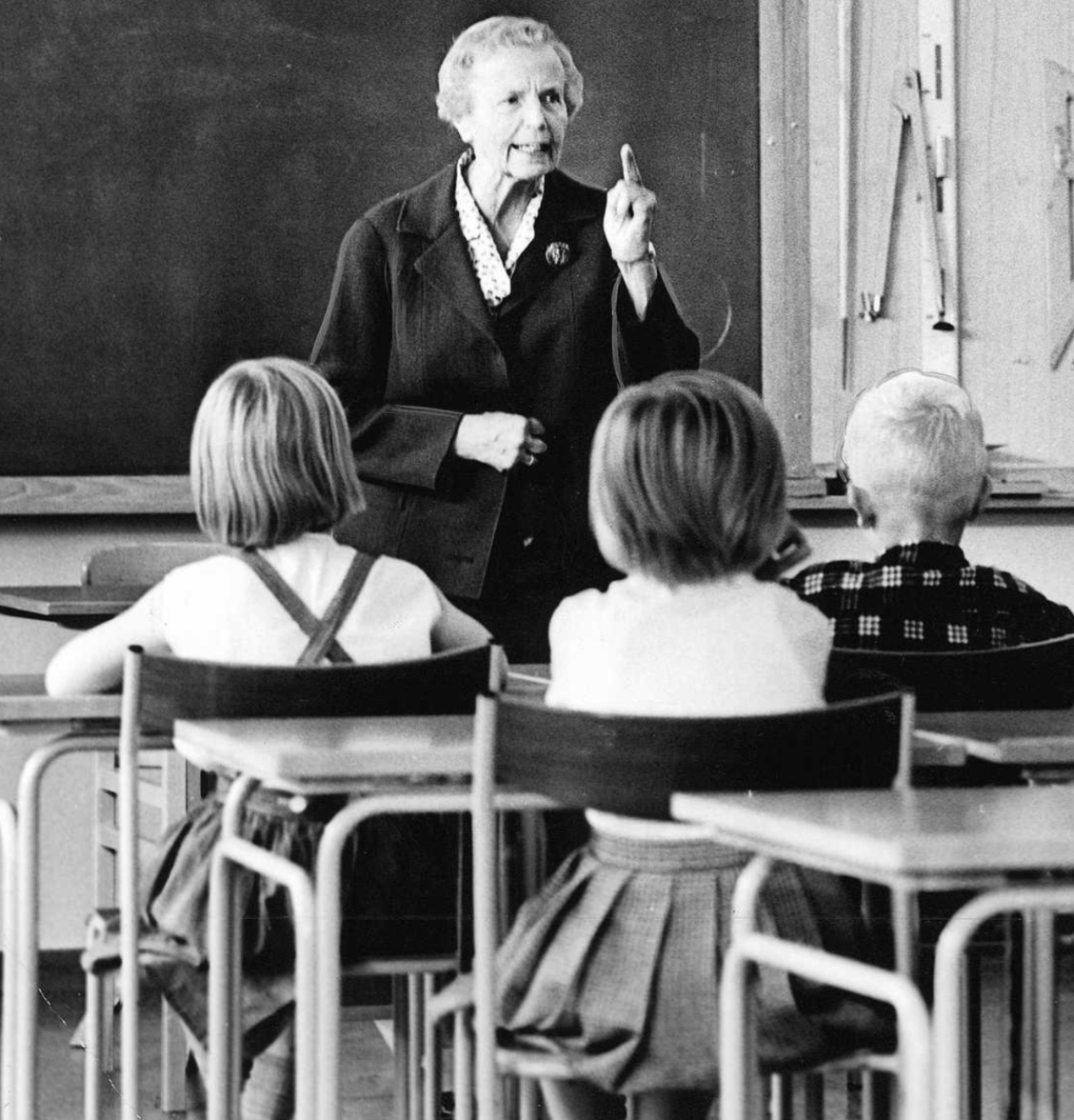En äldre kvinna undervisar en grupp skolbarn i ett klassrum med en svart tavla i bakgrunden