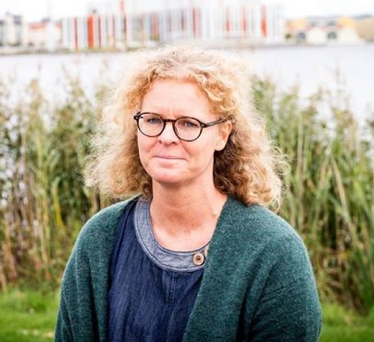 Person med lockigt hår, grön kofta och glasögon tittar in i kameran. 