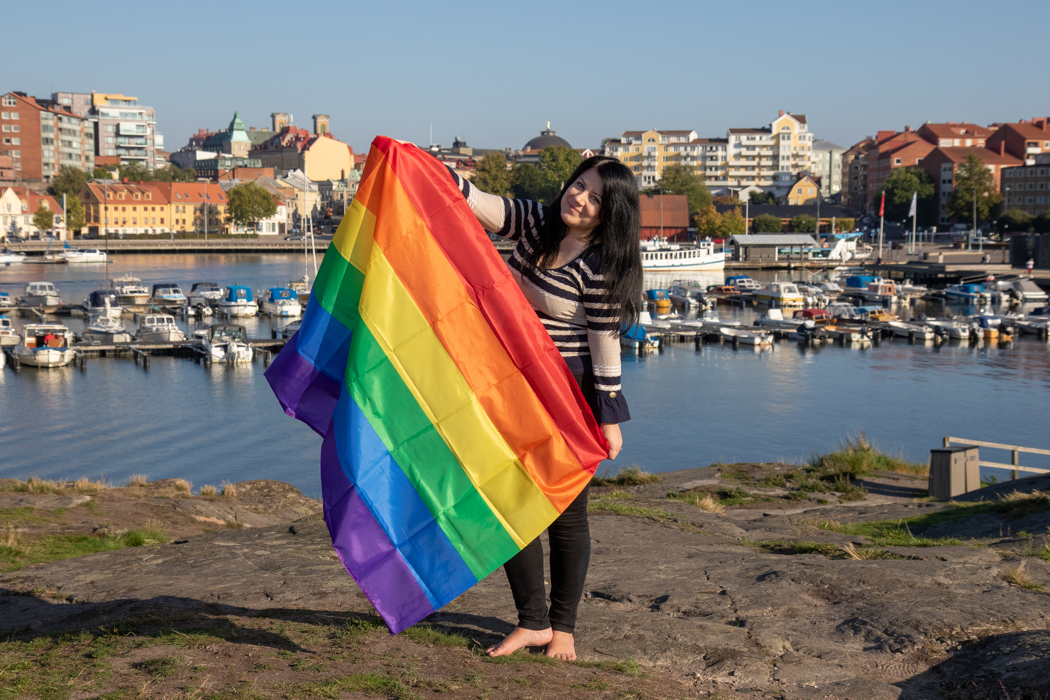 Sophia Ahlin står barfota på en klippa vid vattnet och håller upp en stor regnbågsflagga framför sig.