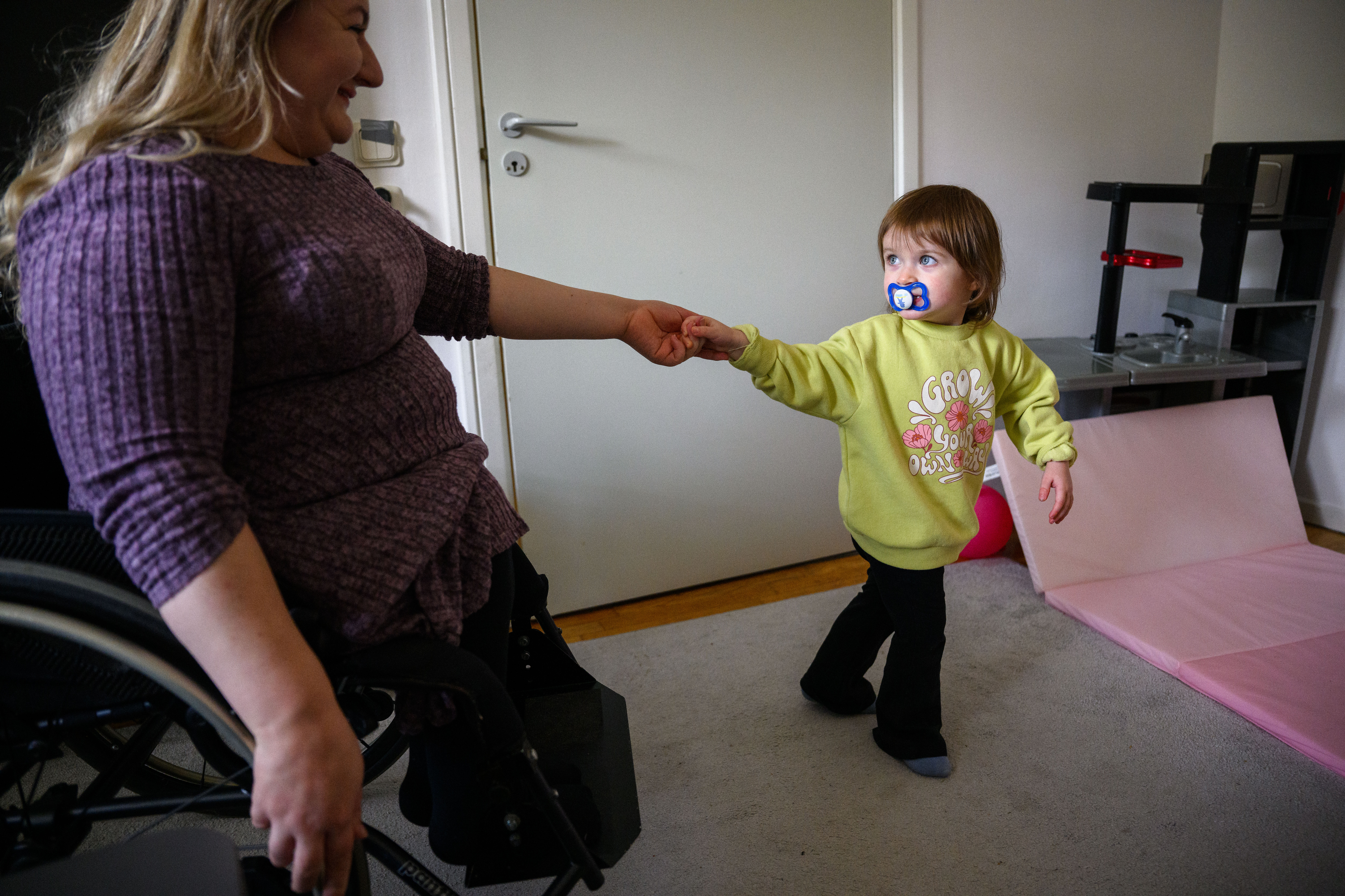 En mamma som sitter i rullstol håller ett litet barn i handen och ler mot barnet. Barnet har en napp i munnen och tittar tillbaka på mamman 