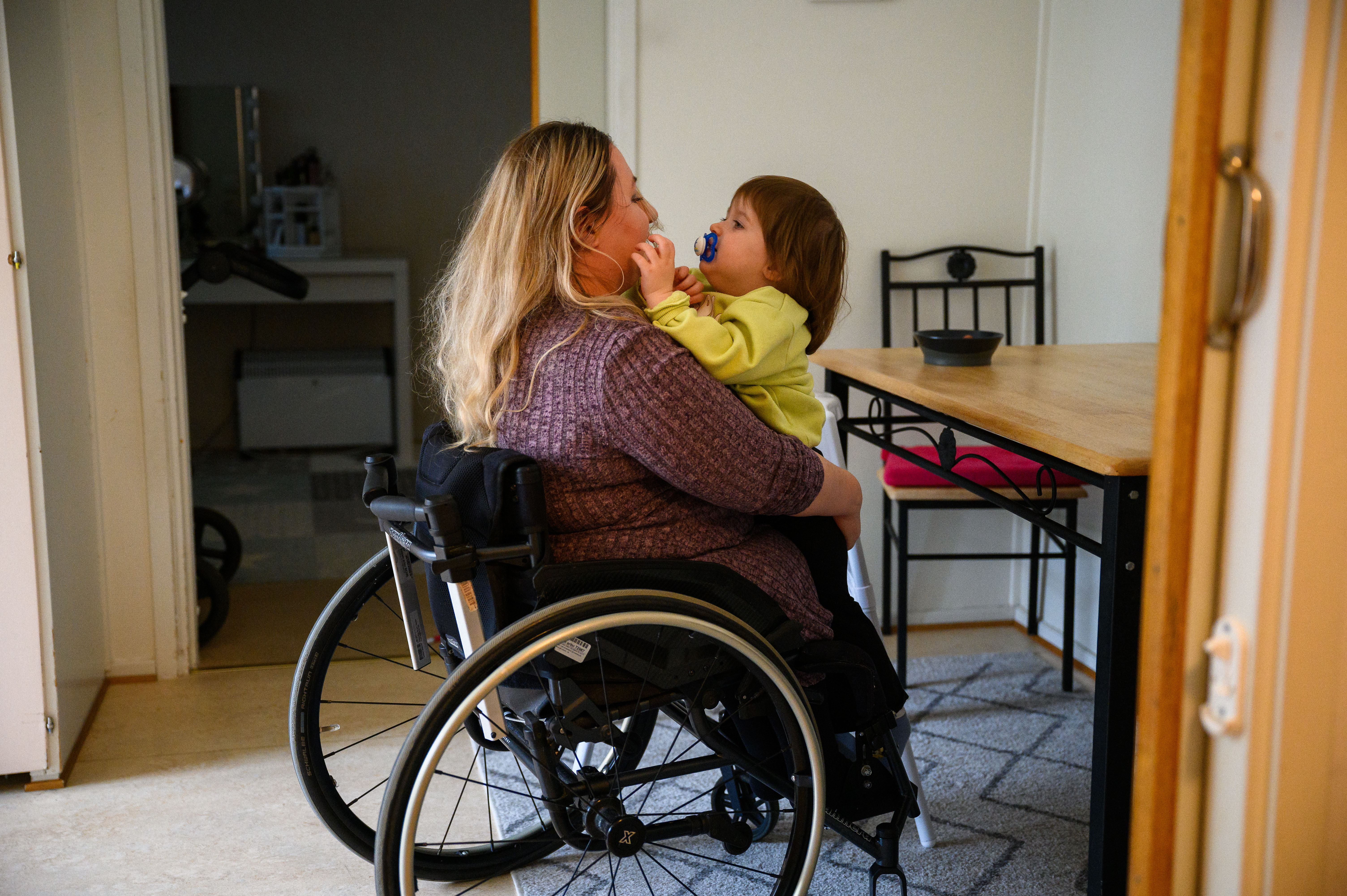 En mamma som sitter i rullstol och har ett barn i knäet. Barnet och mamman kramas och tittar på varandra. 
