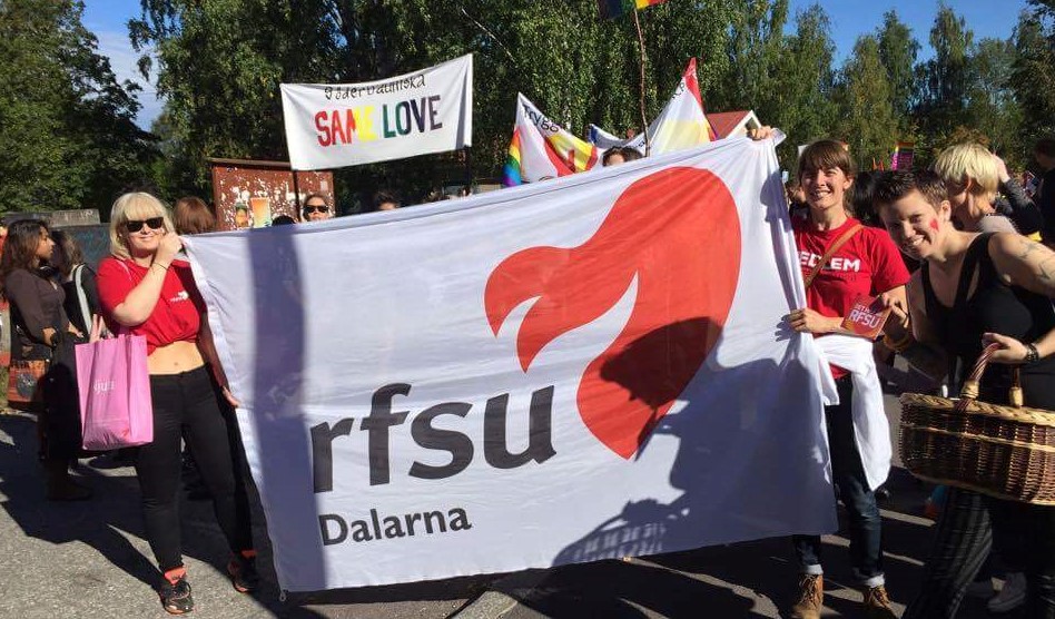 Två personer i röda t-shirts står och håller en vit banderoll med RFSU Dalarnas logotyp. Folk står bakom dem och runtomkring dem. 