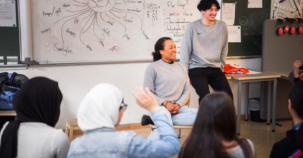 En person sitter och en annan står framför en whiteboard tavla. Framför dem sitter ungdomar på stolar och tittar mot dem.