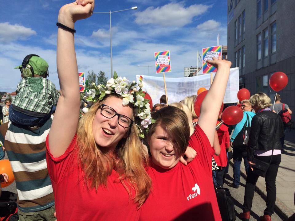 Två personer i röda t-shirts håller varandra om ryggen, ler mot kameran och lyfter ena armen upp i luften. I bakgrunden syns marscherande människor som i en parad.