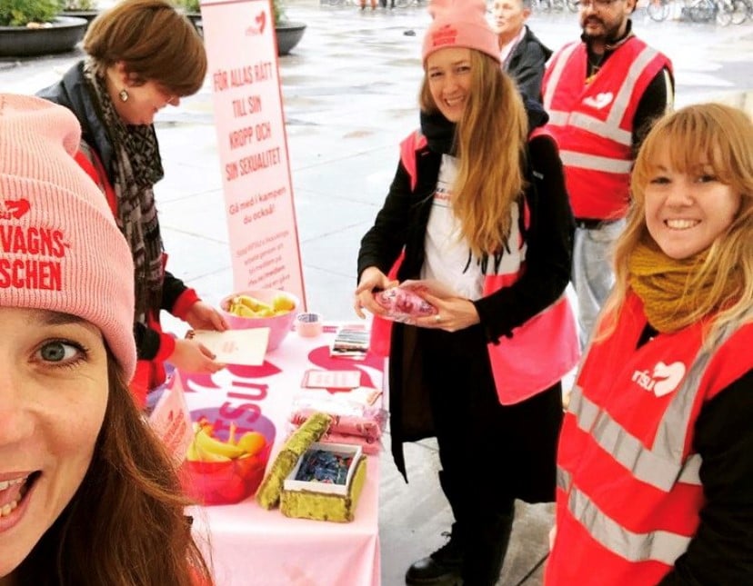 Fem personer i röda och rosa RFSU-västar står vid en RFSU monter utomhus.