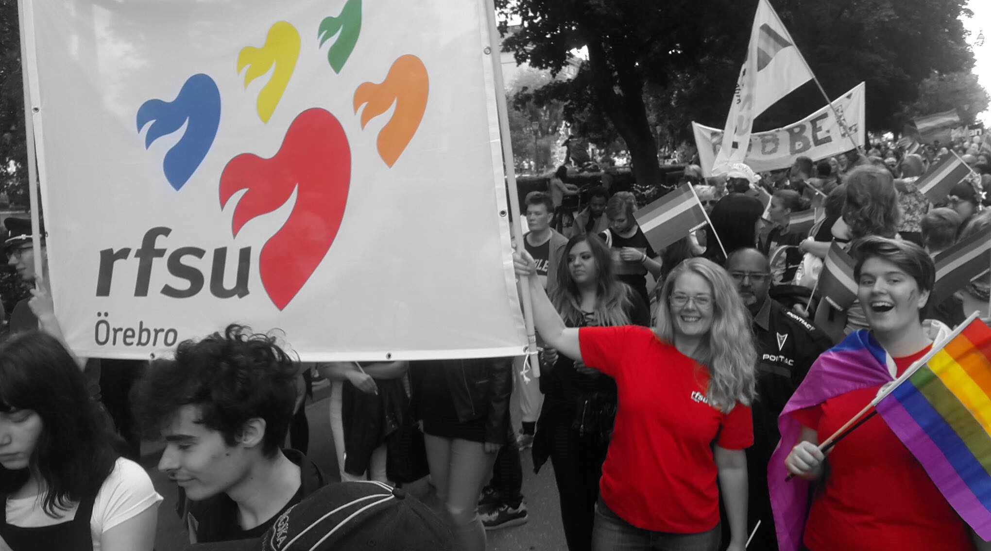 Svartvit bild på ett pride-tåg där endast en pride-flagga, RFSU-banderollen och två röda RFSU t-shirts är i färg.