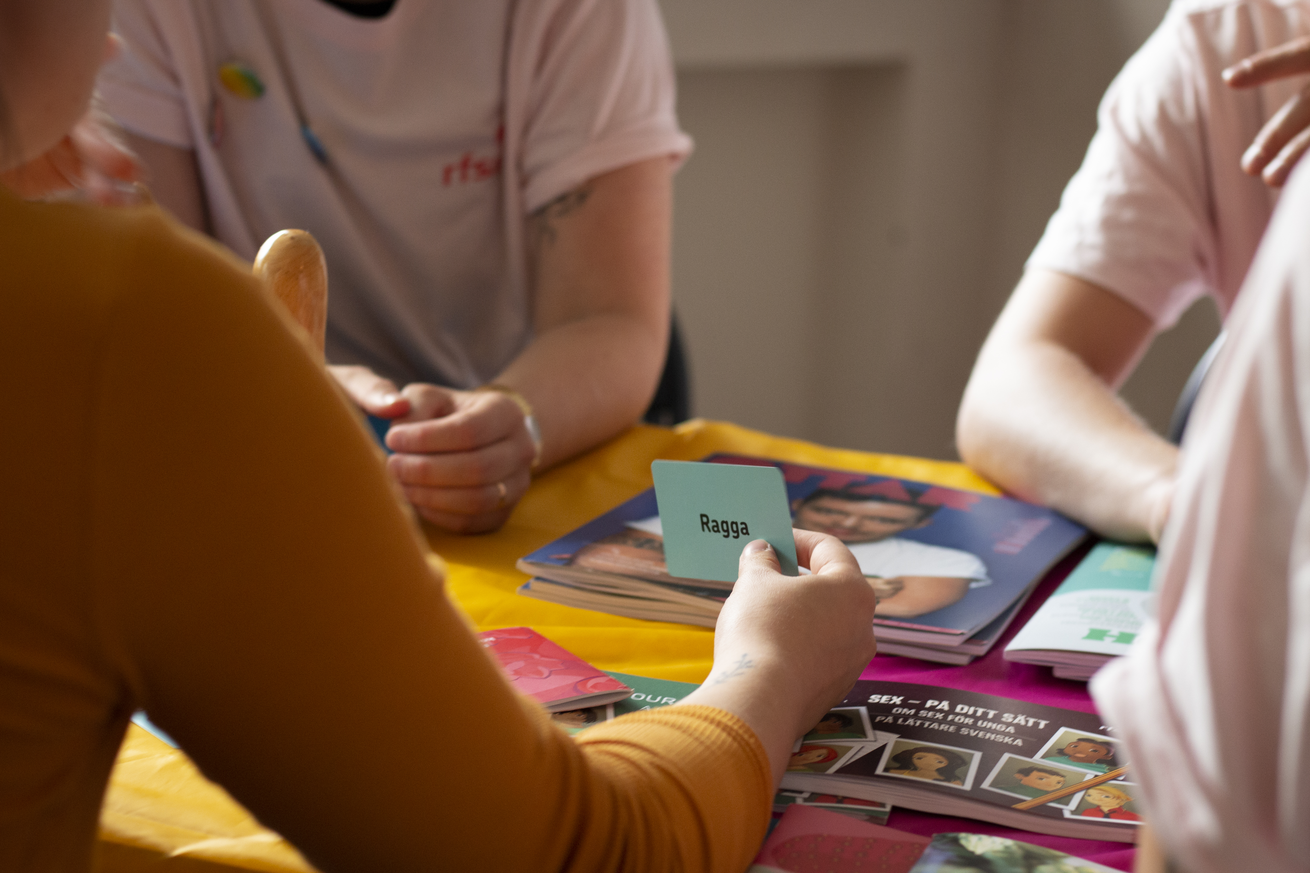 Fyra personer sitter vid ett bord och spelar ett kortspel. Endast händerna och överkropparna syns i bild.