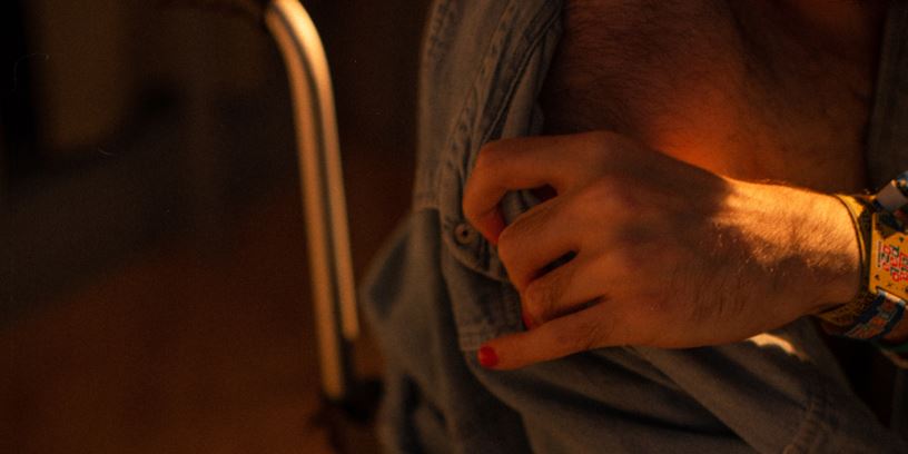 En person sitter i rullstol bredvid en säng. En hand med målade naglar drar av personens skjorta. 